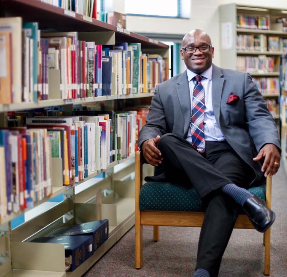 A man in a suit sits smiling on a chair between bookshelves in a library.
